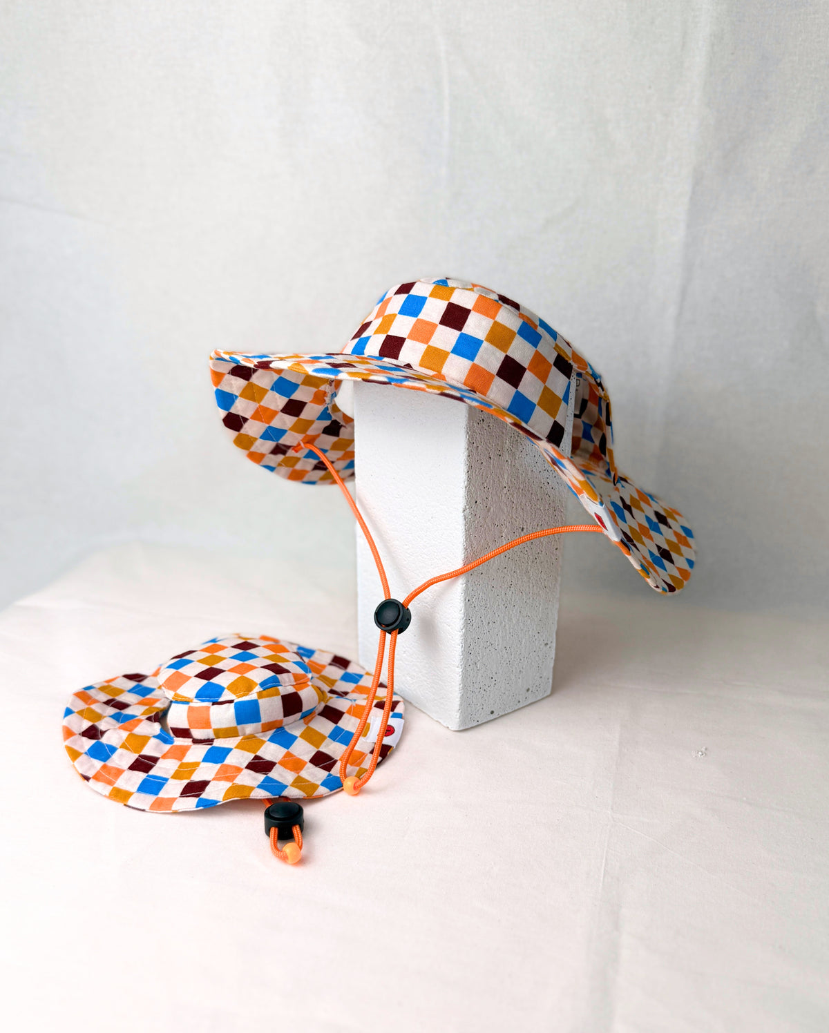 Two colorful diamond-patterned dog sun hats with an orange chin strap, sitting against a plain white backdrop.