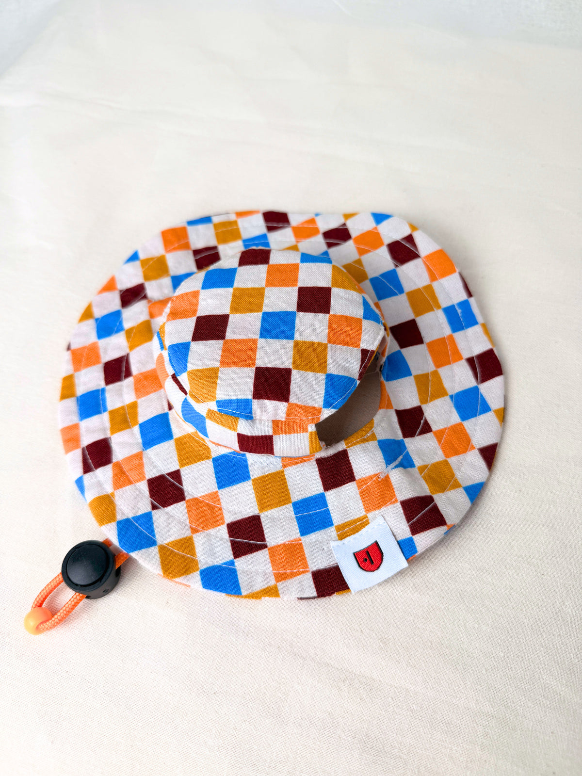 A colorful diamond-patterned dog sun hat with an orange chin strap, sitting against a plain white backdrop.