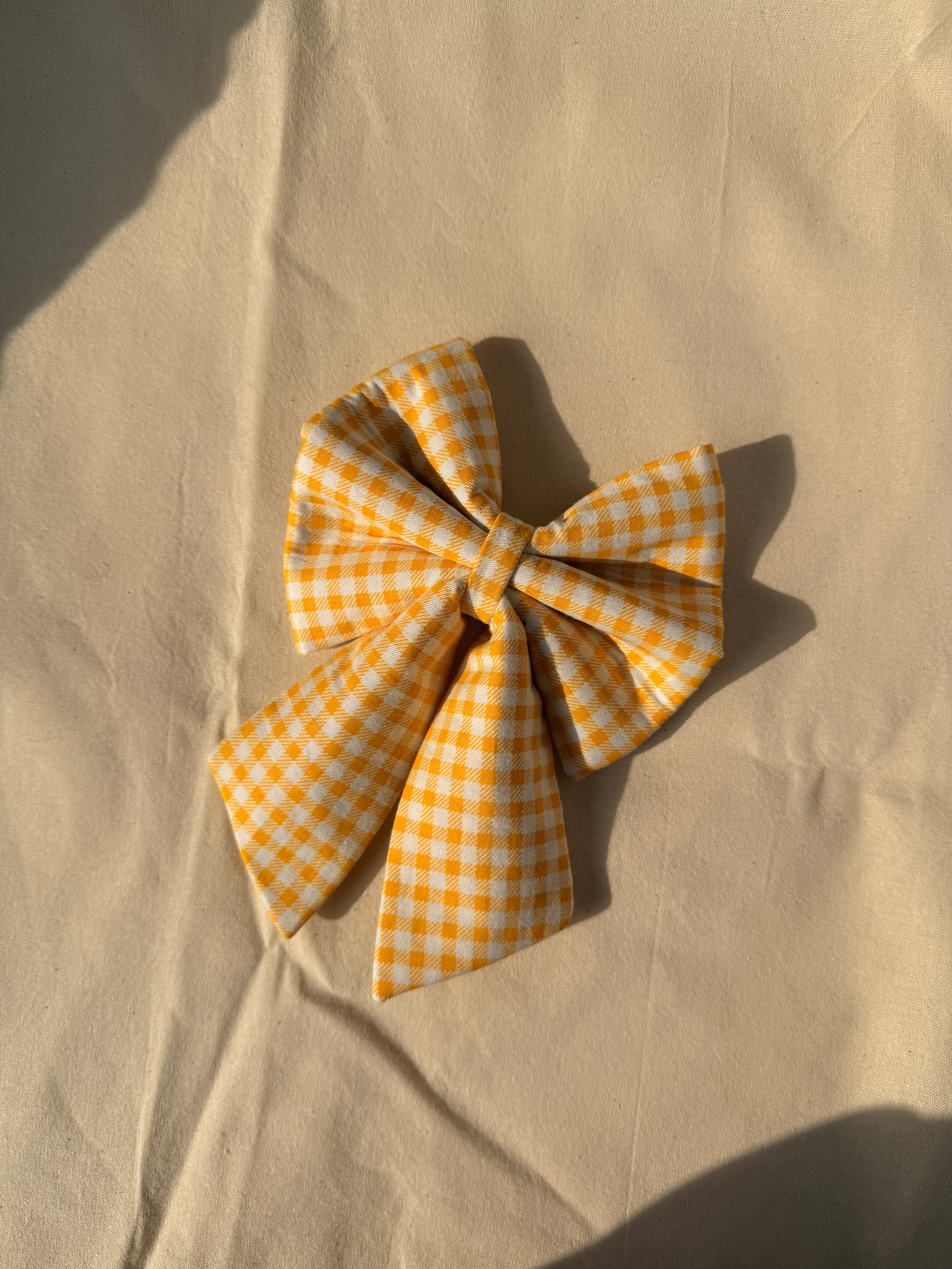 A yellow and white gingham bow accessory, styled on a beige fabric background with warm natural sunlight and shadows.