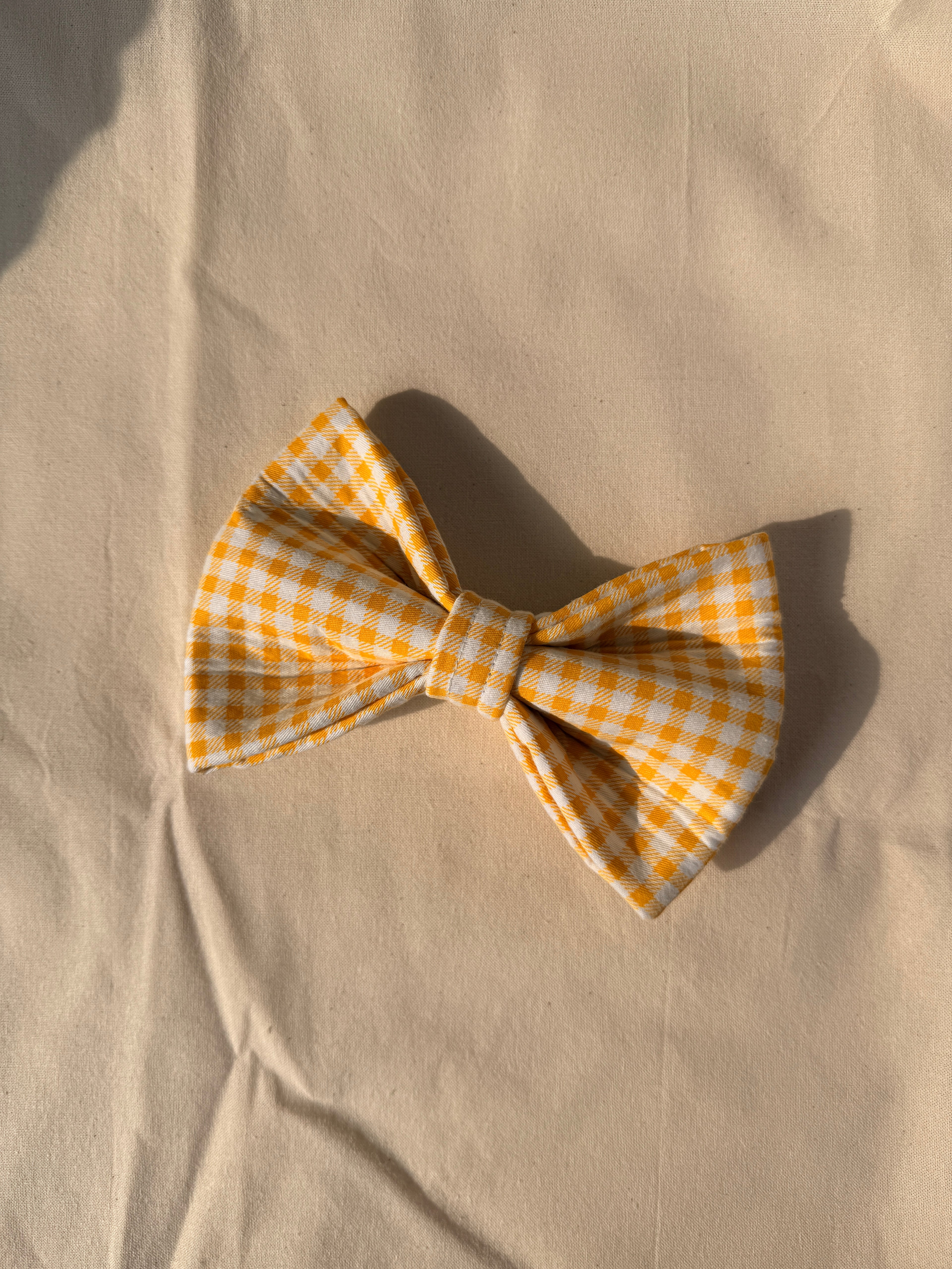 A yellow and white gingham bow tie laid flat on a beige fabric background with a soft shadow.