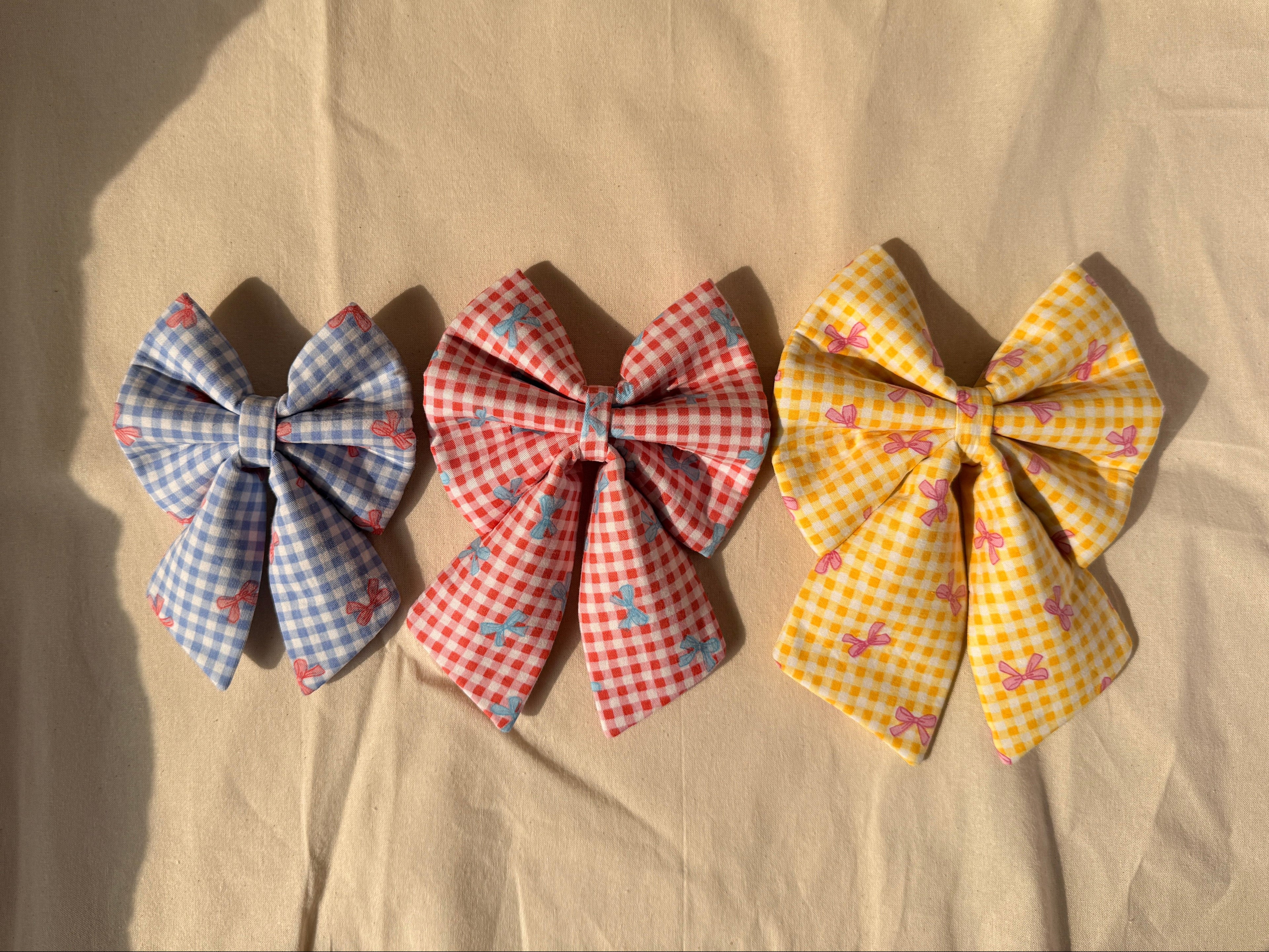 Three fabric hair bows arranged vertically on a beige fabric background. The top bow is light blue and white gingham with small pink flamingos.