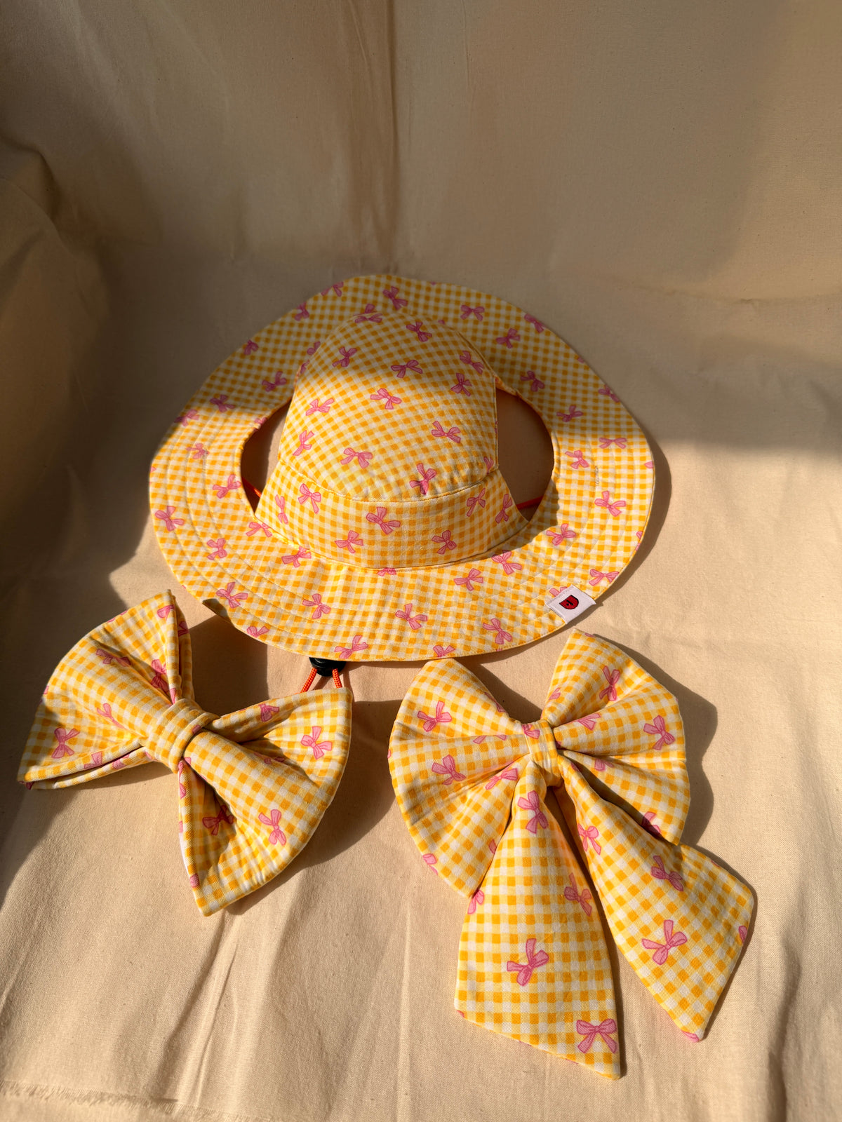 A yellow and white gingham dog sun hat with pink bow patterns, displayed with two matching fabric bow tie and sailor bow on a beige fabric background.