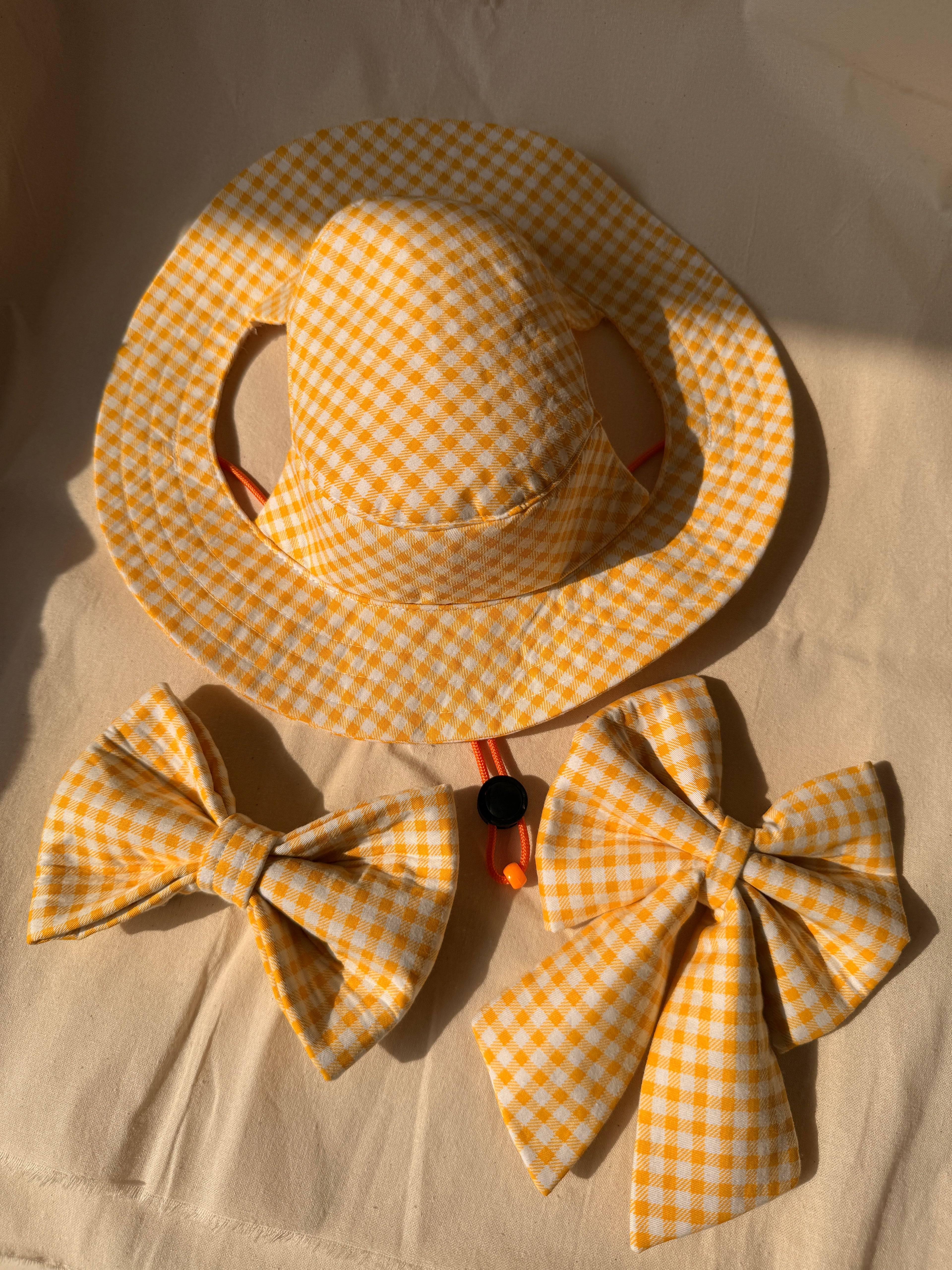A yellow and white gingham sun hat with matching bow accessories, laid flat on a beige fabric background.