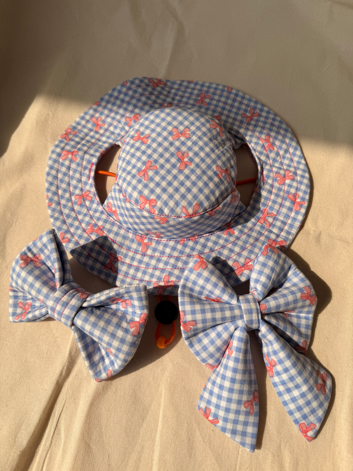 A blue and white gingham dog sun hat with two matching fabric bows, all featuring a small pink bow print, displayed on a beige fabric background.