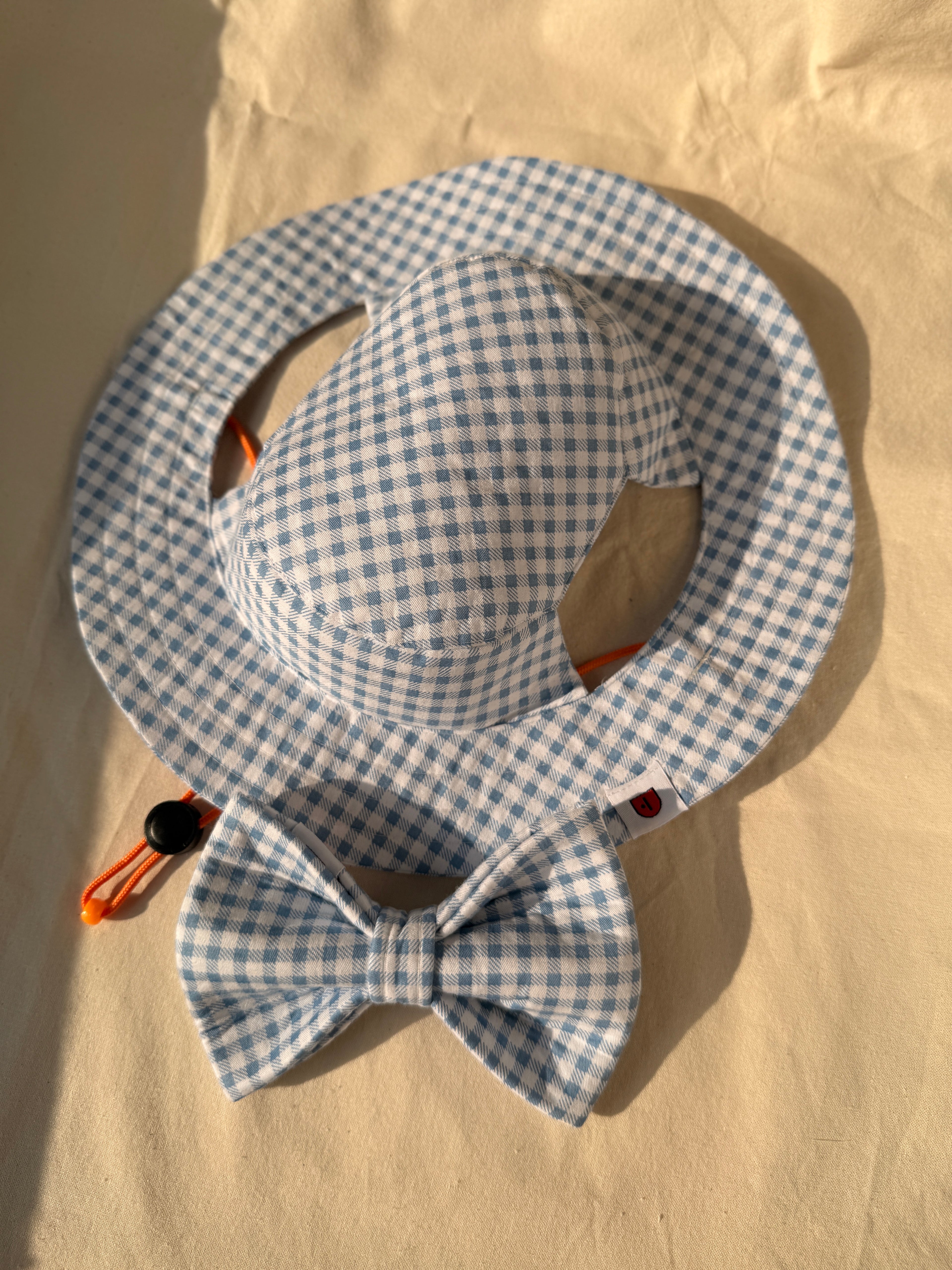 A light blue and white gingham sun hat with a matching bow tie, laid flat on a beige fabric background.